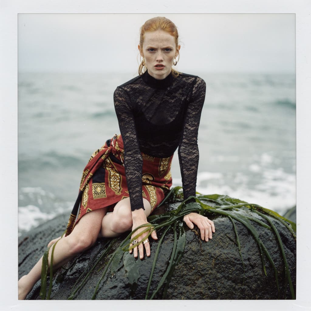 AI generated: a woman sitting on top of a rock next to the ocean, black turtleneck lace dress, lara stone, layout,...