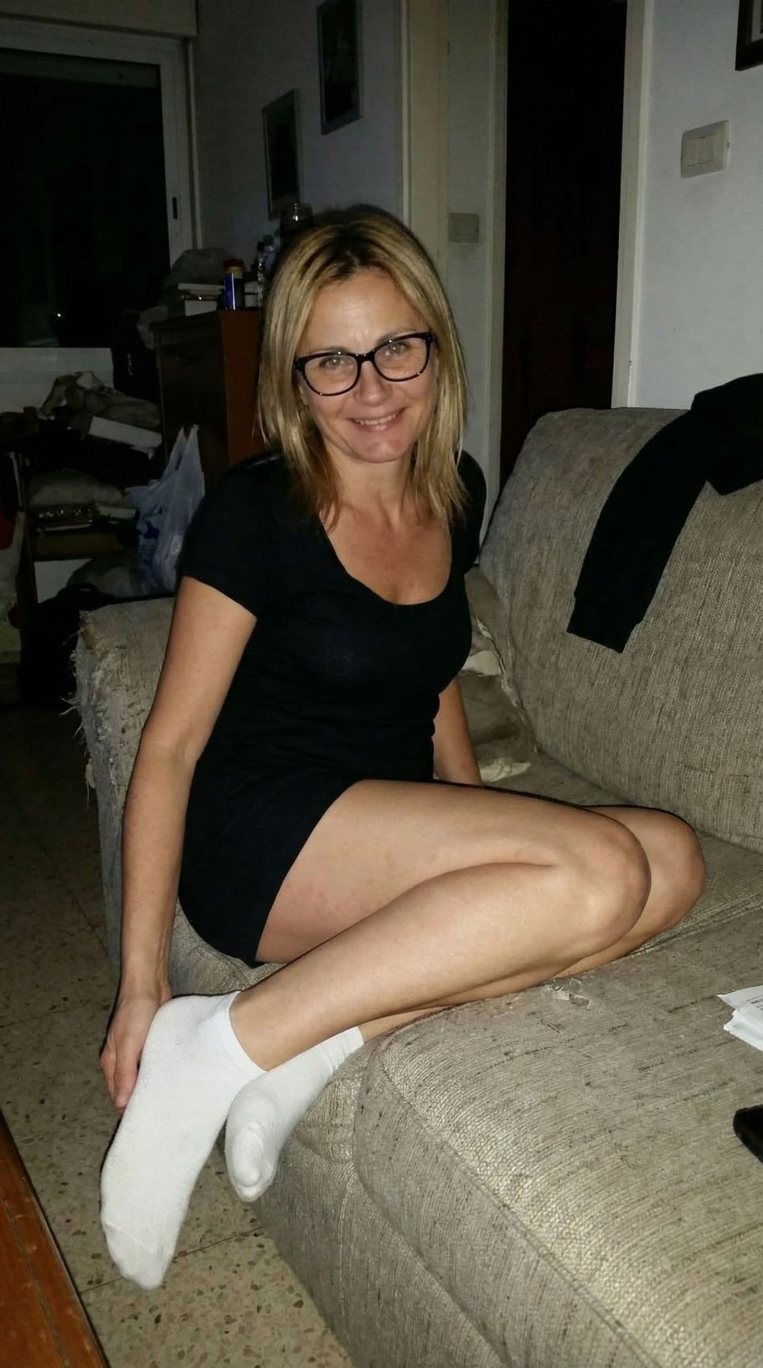 AI generated: Cute, looking at me,a sloppy snapshot. Sitting on a couch at night teasing, mini top and white socks...