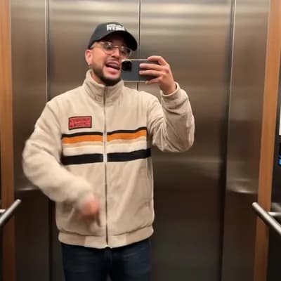 AI generated video: A man is standing in an elevator taking a mirror selfie. He is wearing a light beige corduroy zip-up...