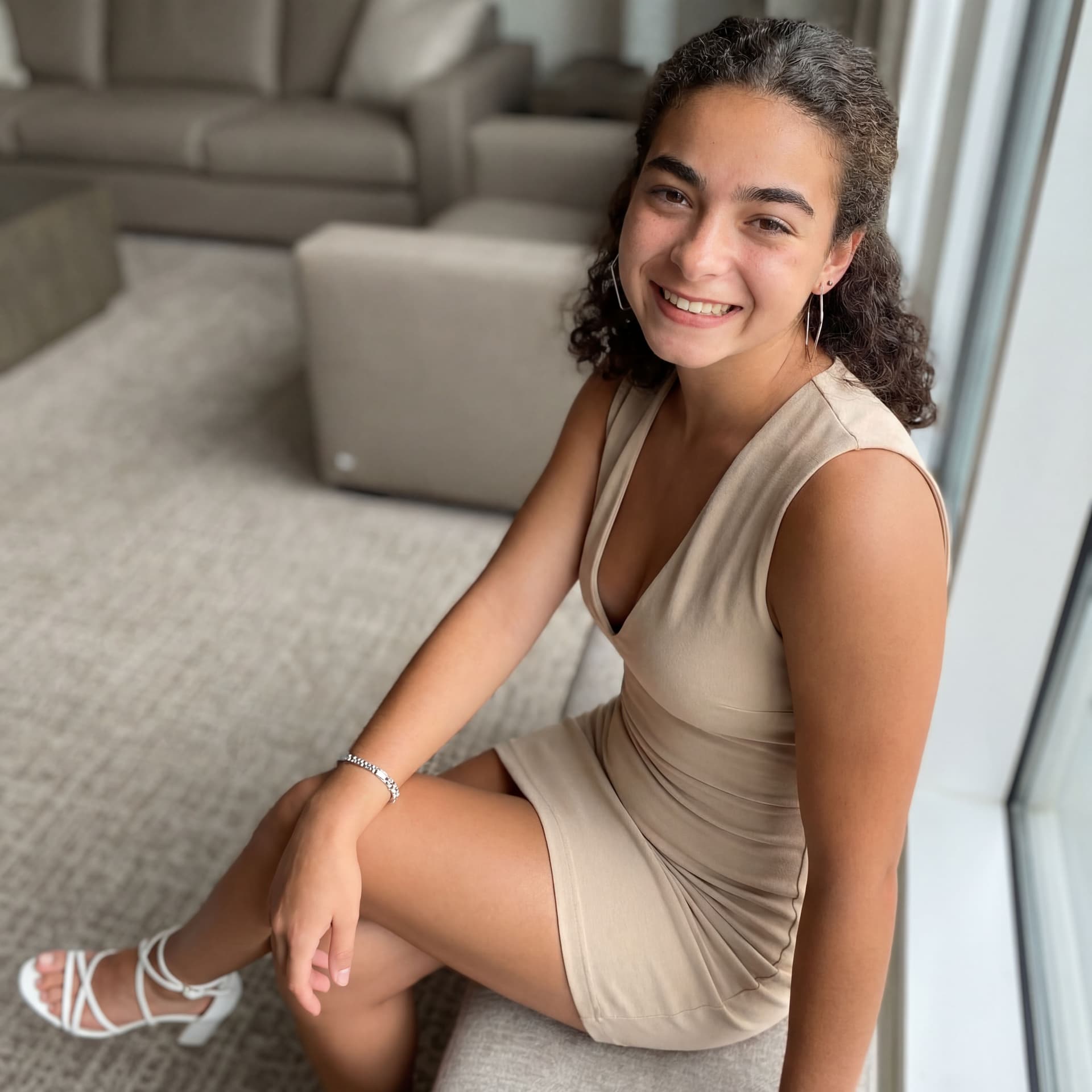 AI generated: warm, inviting smile. She wears a form-fitting, sleeveless beige mini-dress with a deep V-neckline, ...
