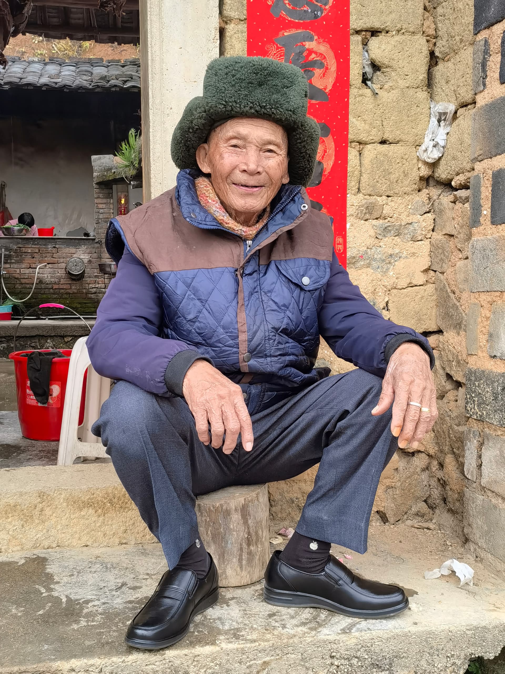 AI generated: 一个中国乡村老人，坐在自己的老房子的台阶上，带着军大帽，穿着羽绒服，慈祥的笑脸