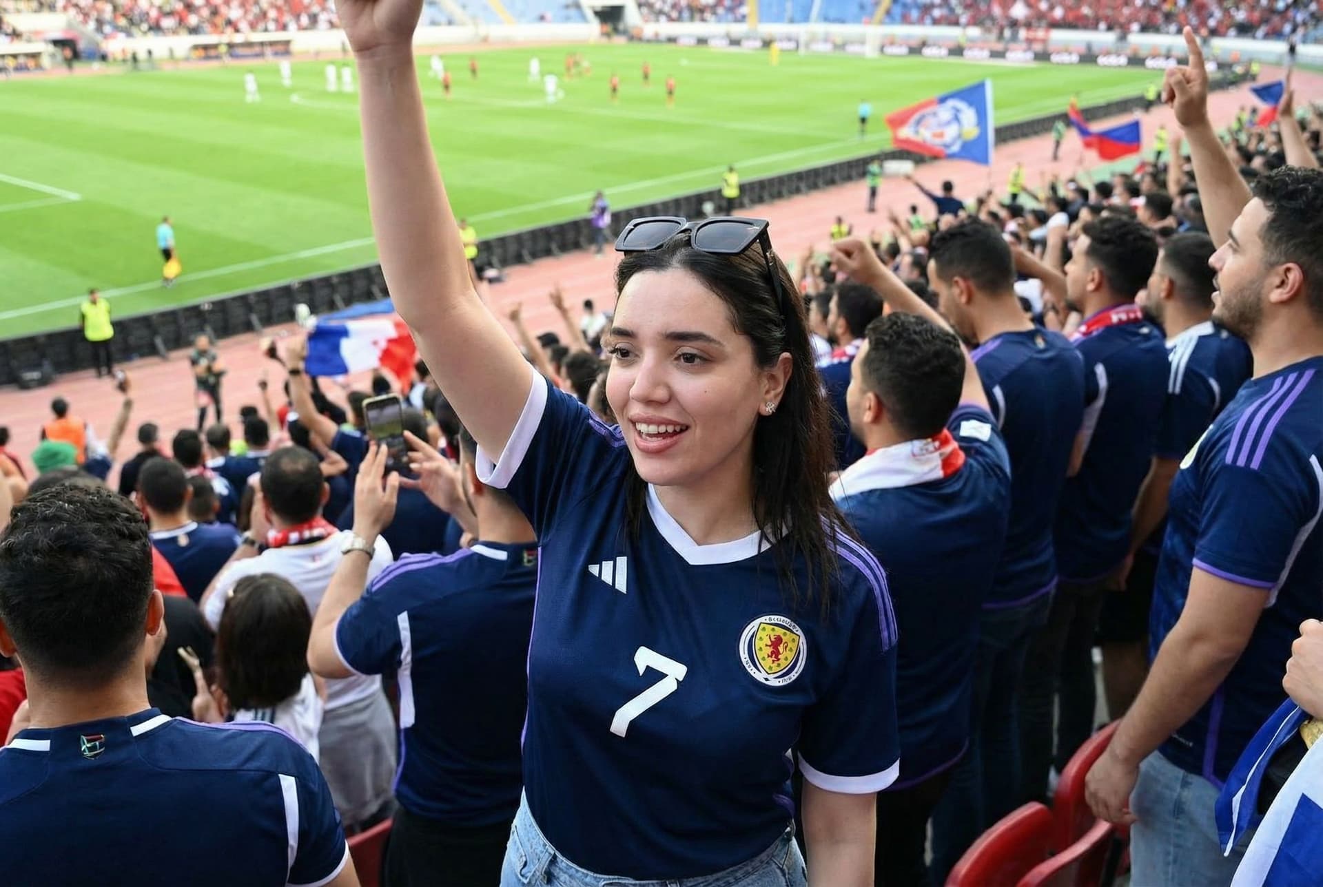 AI generated: change the scenario, put her in the crowd with a football team shirt watching a match