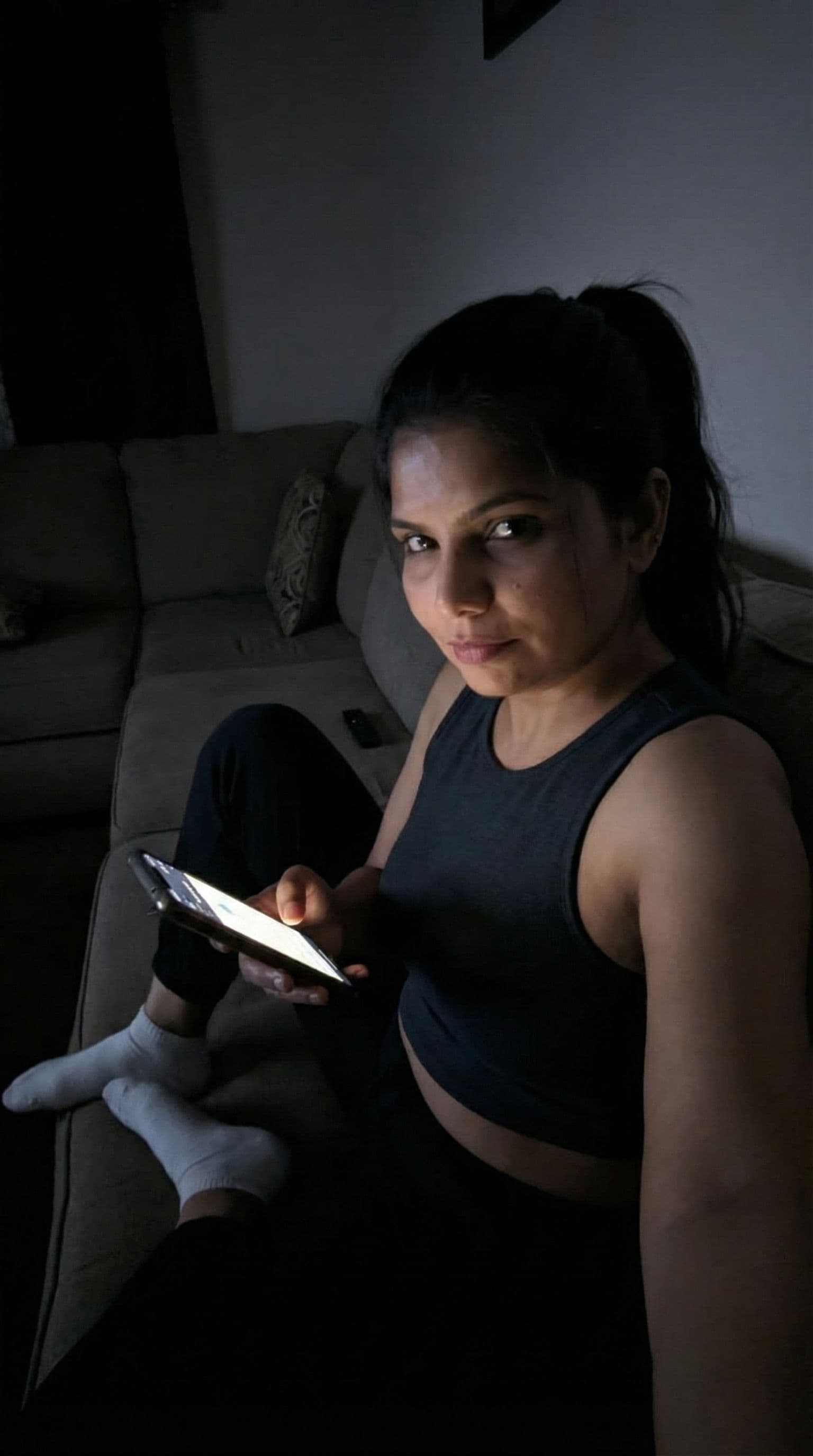 AI generated: Indian, Taking a selfie on a couch at night, no lights, wearing a cropped home tanktop and white soc...