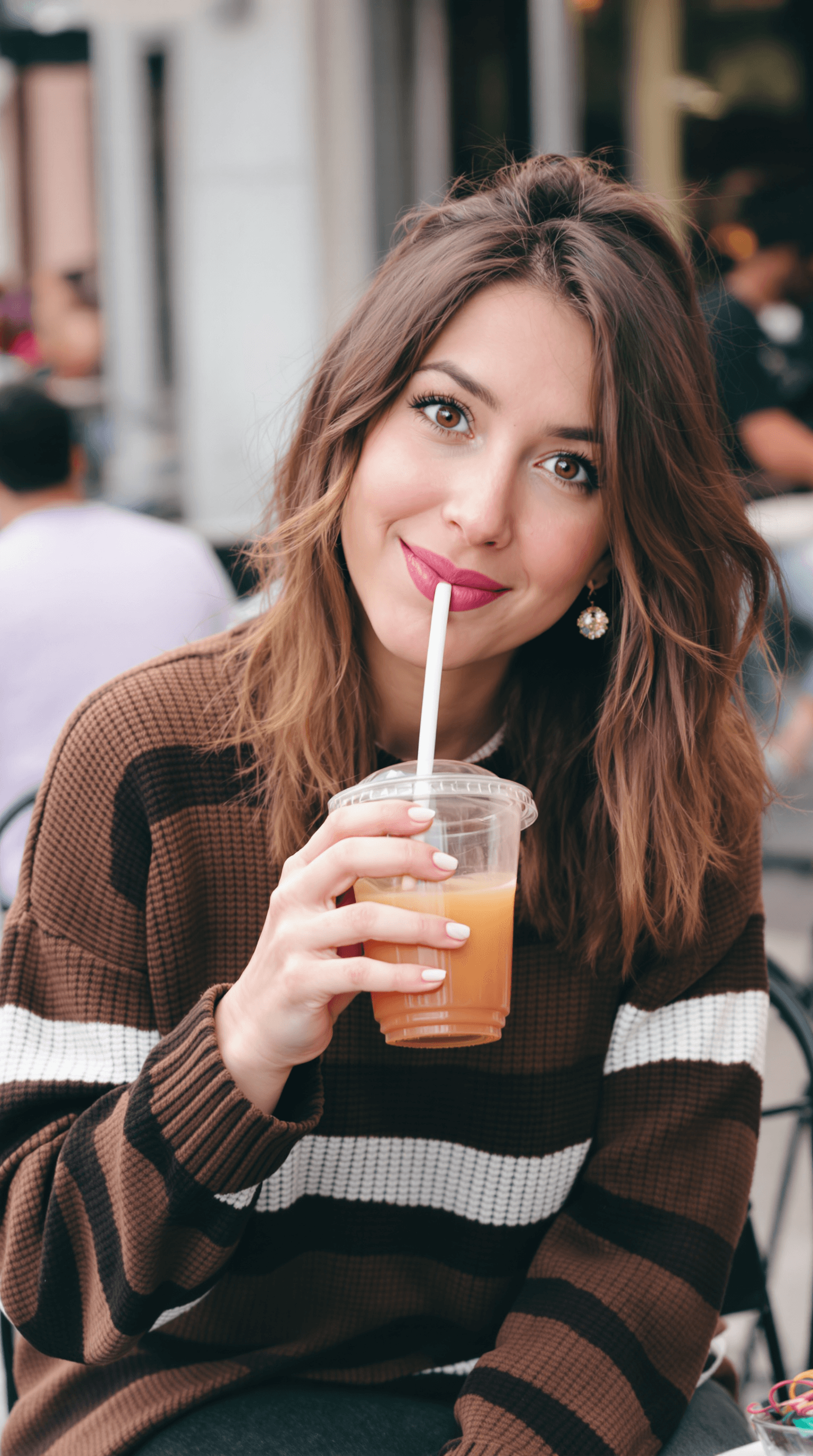AI generated: A woman sitting outdoors, sipping a drink from a transparent cup with a straw, wearing a brown strip...